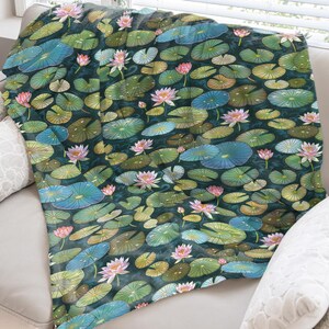 May include: A throw blanket featuring a watercolor design of lily pads and pink water lilies. The blanket has a dark teal background with green, blue, and yellow lily pads. The throw is draped over a white sofa with white pillows.