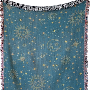 Puede incluir: Una manta tejida azul turquesa con un patrón celestial con soles, lunas y estrellas doradas. La manta tiene un borde con flecos.