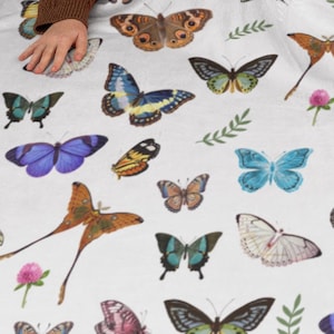 May include: A white throw blanket with a colorful butterfly pattern. The blanket features a variety of butterfly species in shades of blue, green, yellow, orange, and brown. The butterflies are arranged in a random pattern on the blanket.