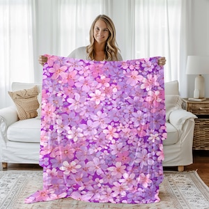 May include: A floral blanket featuring a pattern of pink and purple flowers on a deep purple background. The blanket is held up, showcasing the full design. The flowers are densely packed, creating a vibrant and colorful textile.