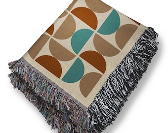 May include: Folded throw blanket with a geometric pattern of brown, teal, and tan semi-circles. The blanket has a fringed edge with a mix of colors, including black, brown, and blue. The blanket is made of a soft material.