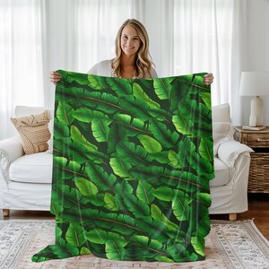 May include: A large, rectangular blanket featuring a vibrant pattern of overlapping green banana leaves. The leaves have varying shades of green with visible veins and a dark background. The blanket is held up, showcasing its full design.