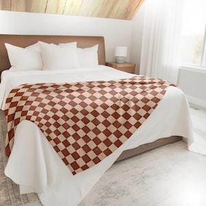 May include: A white bed with a brown and cream checkered throw blanket. The blanket is folded in a triangle shape at the foot of the bed.