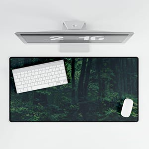 Puede incluir: Una escena de bosque verde oscuro con un teclado blanco y un ratón blanco sobre una alfombrilla de escritorio negra. La alfombrilla está diseñada para parecer un bosque con árboles y hojas.
