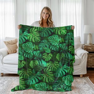 May include: A green blanket with a repeating pattern of large, tropical monstera leaves. The leaves are various shades of green, with detailed veining, set against a dark background. The blanket is held up, showcasing the design.