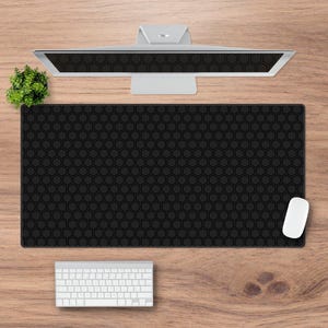 Op de afbeelding: Een zwarte muismat met honingraatpatroon met een witte computermuis op een houten bureau. Een wit toetsenbord en een computermonitor zijn ook zichtbaar.