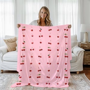 May include: A pink blanket featuring a pattern of red cherries with green leaves. The cherries are evenly spaced across the blanket's surface. The blanket is held up in front of a white couch and curtains.