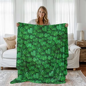 May include: A green blanket featuring a repeating pattern of monstera leaves. The blanket is held up, showcasing the vibrant green color and the detailed leaf design. The blanket is made of a soft material and is perfect for adding a touch of nature to any room.