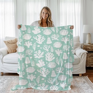 May include: A soft, light blue throw blanket with a white coral reef pattern. The blanket is being held up by a person in a white room.