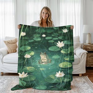 May include: A green frog sits on a lily pad in a pond with white water lilies. The pond is surrounded by green lily pads and leaves. The background is a blurry image of a person holding the blanket.