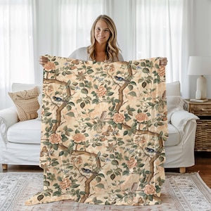 May include: A cream-colored throw blanket with a floral pattern featuring pink roses, green leaves, and blue birds. The blanket has a vintage aesthetic and is perfect for adding a touch of elegance to any room.