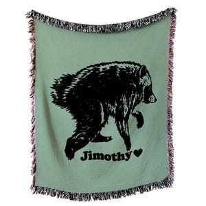 May include: A sage green woven throw blanket featuring a black graphic of a bear walking. The text "Jimothy" is printed in black below the bear, followed by a small black heart. The blanket has a decorative fringe along all four edges.