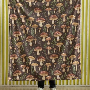 May include: A brown fleece throw blanket with a repeating pattern of mushrooms and leaves. The blanket is being held up by a person, but the person's hands are not visible in the image.