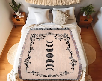 May include: A woven throw blanket with a cream background and black floral design. The center of the blanket features a black design of a moon in various phases.