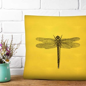 May include: A square throw pillow in a vibrant yellow color featuring a detailed black line art illustration of a dragonfly centered on the front. The dragonfly has intricate wing patterns and a segmented body, providing a high contrast design.