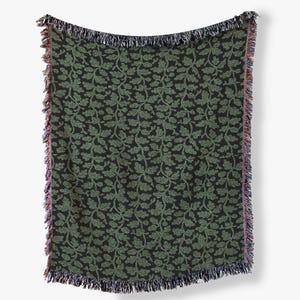 May include: A woven throw blanket featuring a repeating pattern of green oak leaves and acorns set against a solid black background. The rectangular textile has decorative fringed edges along the top and bottom, showcasing a blend of multicolored threads.