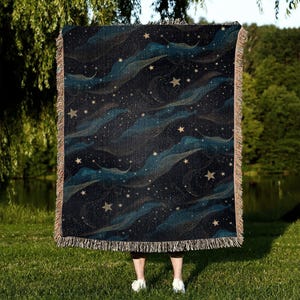 Puede incluir: Una manta tejida con un fondo azul oscuro y un patrón de estrellas doradas y líneas en espiral. La manta tiene un borde con flecos.