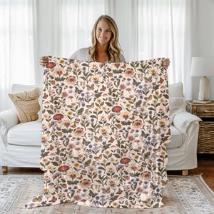 May include: A cream-colored throw blanket with a floral pattern featuring butterflies, mushrooms, and flowers in shades of pink, red, orange, yellow, green, and blue.