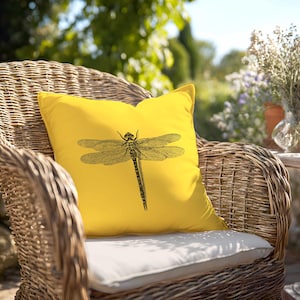 May include: A square throw pillow in a vibrant yellow color featuring a detailed black line art illustration of a dragonfly centered on the front. The pillow has a smooth fabric texture and is positioned upright on a woven wicker chair.