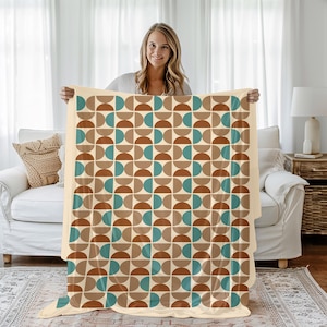 May include: A beige blanket with a repeating geometric pattern of brown, teal, and tan semi-circles. The blanket is held up, showcasing its design and soft texture. The blanket is a cozy home decor item.
