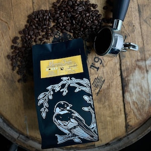 May include: A black coffee bag with a white bird illustration and the text "Mexican Chiapas" and "Snowbird Mountain Coffee Co."  The bag is sitting on a wooden surface with coffee beans and a silver coffee tamper.