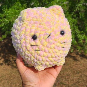 May include: A crocheted plush toy shaped like a cat with pink and yellow yarn. The toy has black eyes and a black stitched nose and mouth.