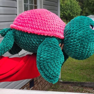 May include: A crocheted stuffed turtle toy with a pink shell and green body. The turtle has a large head with a single eye.
