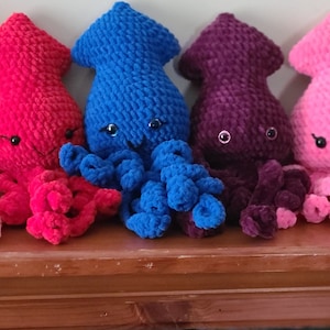 May include: Five crocheted plush squid toys in different colors: pink, red, blue, purple, and pink. Each squid has a smiling face with black eyes and a small mouth.