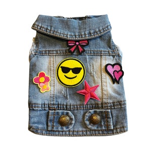May include: A light blue denim vest with various patches, including a yellow smiley face with sunglasses, a pink flower, a pink star, and a pink heart.
