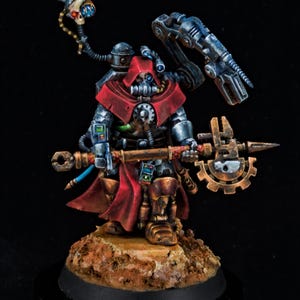 May include: A detailed miniature figure of a character in a red hooded cloak and metallic armour. The figure has a respirator mask and holds a staff with a gear-shaped head and a mechanical arm. A skull with blue eyes is attached to a flexible tube.