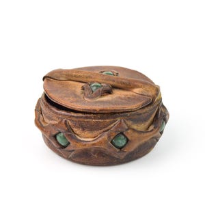 Puede incluir: Una caja redonda de cuero marrón con tapa abisagrada. La caja está adornada con detalles de piedras preciosas verdes y una correa decorativa de cuero. La caja tiene aproximadamente 10 cm de diámetro.