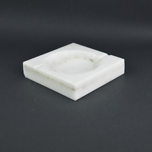 May include: A white marble ashtray with a round depression in the centre. The ashtray is square and has a smooth, polished surface.