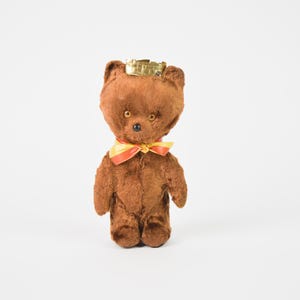 Op de afbeelding: Een vintage bruine teddybeer met een gouden kroon en een geel en rode strik. Het pluche speelgoed heeft kleine, ronde ogen en een zwarte neus. De beer staat rechtop tegen een witte achtergrond.