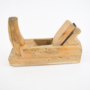 Op de afbeelding: Een vintage houten handvlak, met een gebogen handvat en een mes. Het vlak is gemaakt van lichtgekleurd hout met zichtbare nerf en tekenen van slijtage, wat wijst op de leeftijd en het gebruik. Het mes is donker en contrasteert met het hout.