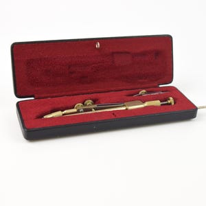 Könnte beinhalten: Ein Vintage-Zeichenzirkel-Set in einem schwarzen Etui mit rotem Samtinterieur. Der Zirkel ist goldfarben mit einem dunkelbraunen Akzent und enthält ein kleines, silberfarbenes Werkzeug. Das Etui ist geöffnet und zeigt die Werkzeuge.