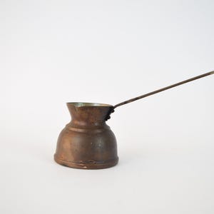 May include: A small, antique copper coffee pot with a long, thin handle. The pot has a rounded body and a slightly flared lip.