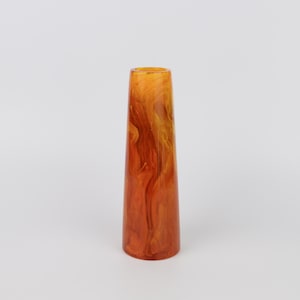 Op de afbeelding: Een lang, cilindrisch object met een uitlopende basis en een smalle opening aan de bovenkant. Het object heeft een wervelend patroon van oranje en gele tinten, waardoor een marmerachtig effect ontstaat. De algemene vorm is taps toelopend en het oppervlak lijkt glad en glanzend.