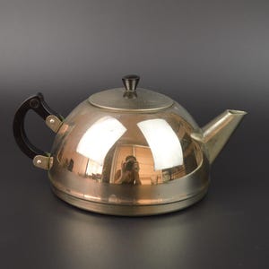 Op de afbeelding: Een zilverkleurige theepot met een afgeronde body, een tuit en een deksel. De handgreep is zwart met zilverkleurige hardware. De theepot reflecteert de omgeving. De theepot wordt waarschijnlijk gebruikt voor het zetten van thee.