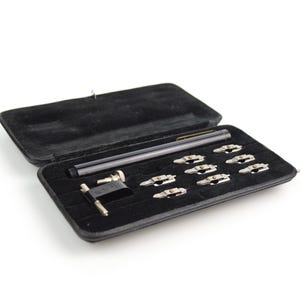 May include: An open black case with a velvet interior, containing a pen, several silver-toned metal objects, and a small tool. The case is rectangular and has a hinged lid. The objects are arranged neatly within the case.