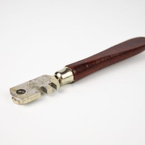 Peut inclure: Un coupe-verre vintage avec un manche en bois brun foncé et une tête de coupe en métal argenté. La poignée est lisse et arrondie, et la tête de coupe comporte une série de petites roues crantées. L'outil est conçu pour couper le verre.