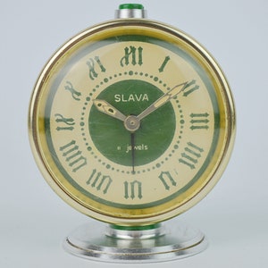 Könnte beinhalten: Ein grüner und goldener Vintage-Wecker mit römischen Ziffern und dem Wort "SLAVA" auf dem Zifferblatt. Der Wecker hat einen grünen Knopf oben und einen silbernen Sockel.