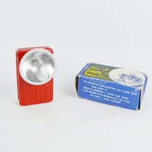 May include: A red flashlight with a white reflector and a blue box with text in Bulgarian. The text on the box reads: "ЕЛ. ФЕНЕРЧЕ ЕДНОЦВЕТНО 2.5 У МОД. 8105 БДС 2643-72 ЦЕНА НА ДРЕБНО 1 БРОЙ 2,80 ЛЕВА сно писмо по 2800.54/30.11.21 год НА OHC ГРАД ПЛЕВОЯ"