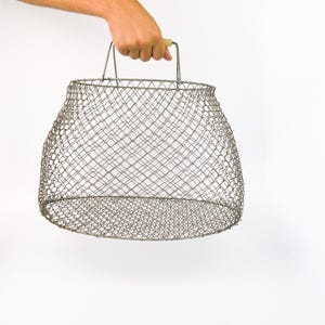 Könnte beinhalten: Ein silberner Drahtgitterkorb mit Holzgriff wird vor weißem Hintergrund gehalten. Der Korb hat eine abgerundete Form. Der Griff ist hellbraun.