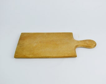 May include: A rectangular wooden cutting board with a handle on the right side. The board is light brown and has a worn, rustic look.