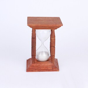 Puede incluir: Un reloj de arena de madera con un bulbo de vidrio transparente y arena blanca. El reloj de arena está sobre una base de madera con tallas decorativas.