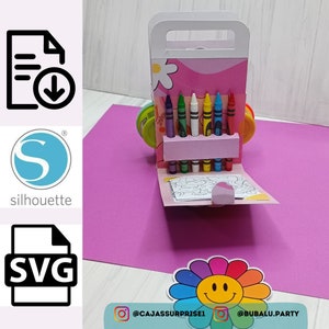 Pode incluir: Uma caixa de cartão rosa e branca com uma alça, decorada com uma flor e um rosto sorridente. A caixa está aberta e revela um conjunto de lápis de cera e uma página para colorir. A caixa está etiquetada com as alças de mídia social @CAJASSURPRISE1 e @BUBALU.PARTY.