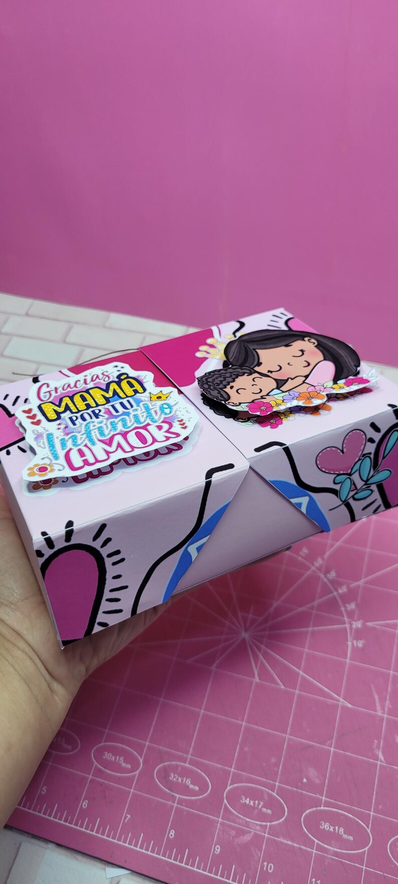 May include: A pink and blue gift box with a colorful design and the text "Gracias Mam&aacute; por tu infinito amor". The box features a cartoon illustration of a mother and child.