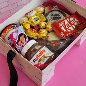 May include: A white gift box filled with various chocolate treats, including Ferrero Rocher, Kit Kat, and Nutella. The box also includes a small bag of breadsticks and a card with a cartoon illustration of a mother and child.