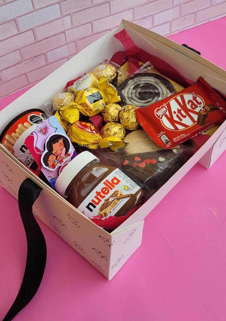 May include: A white gift box filled with various chocolate treats, including Ferrero Rocher, Kit Kat, and Nutella. The box also includes a small bag of breadsticks and a card with a cartoon illustration of a mother and child.