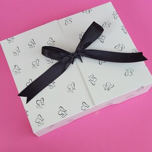 May include: A white gift box with a black ribbon tied in a bow. The box is decorated with a repeating pattern of small hearts.